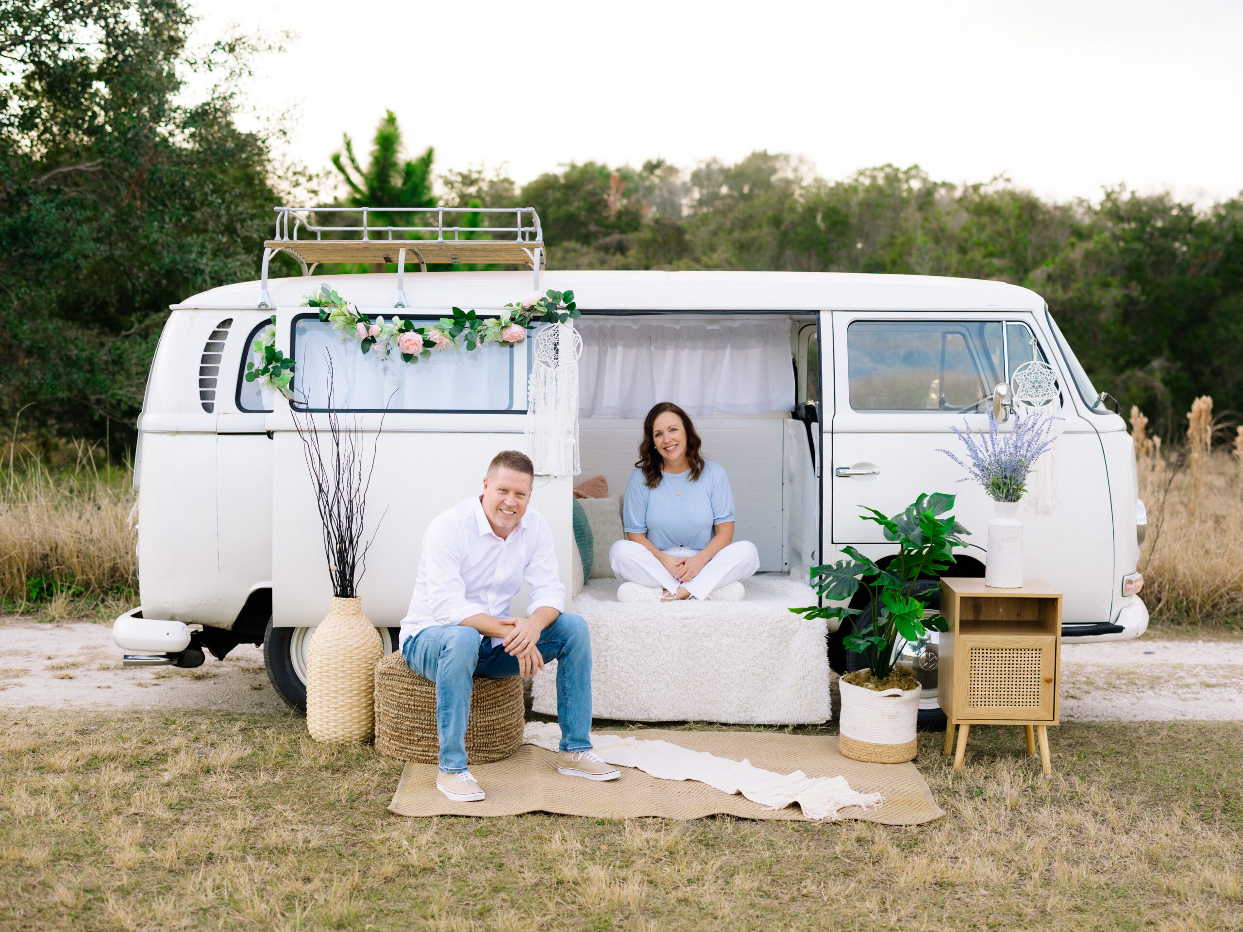 A VW Bus with a couple sitting inside. Thebus is outdoors in a nature location.