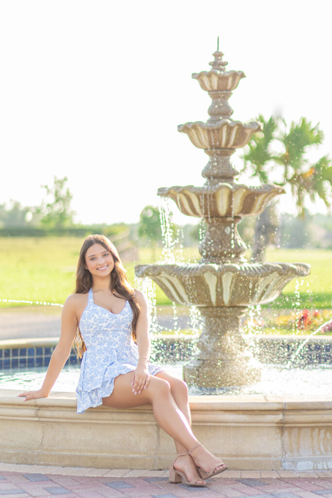 High school senior sitting by a tiered fountain wearing a blue and white romper during her Class of 2025 photo session.