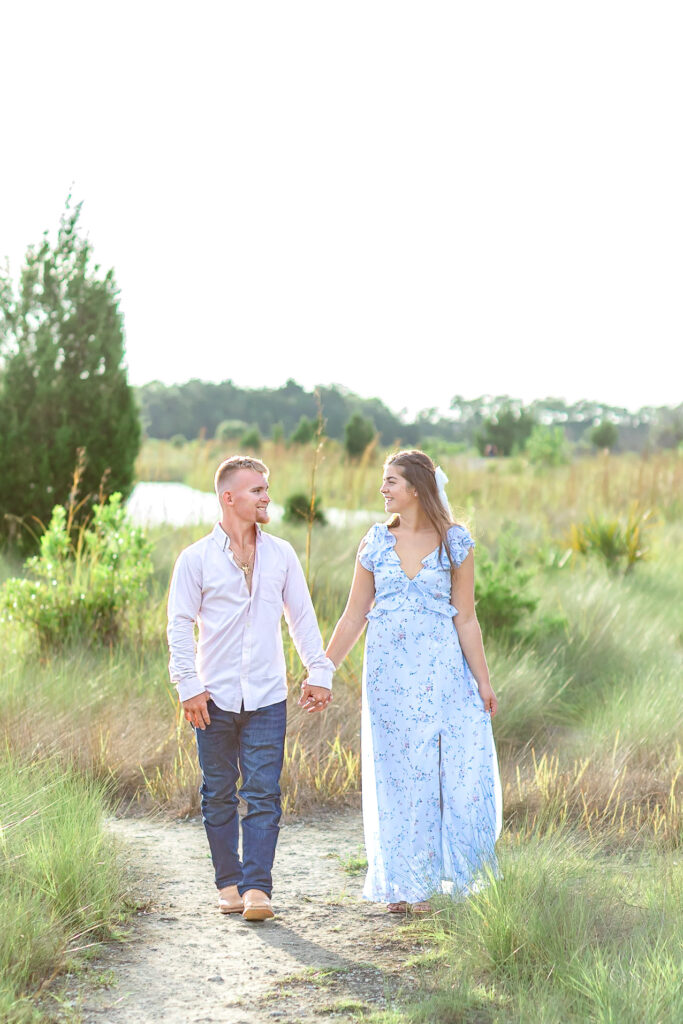 a couple walking on a white trail, surrounded by tall grass. They are dressed up and looking at each other, lovingly,and smiling.