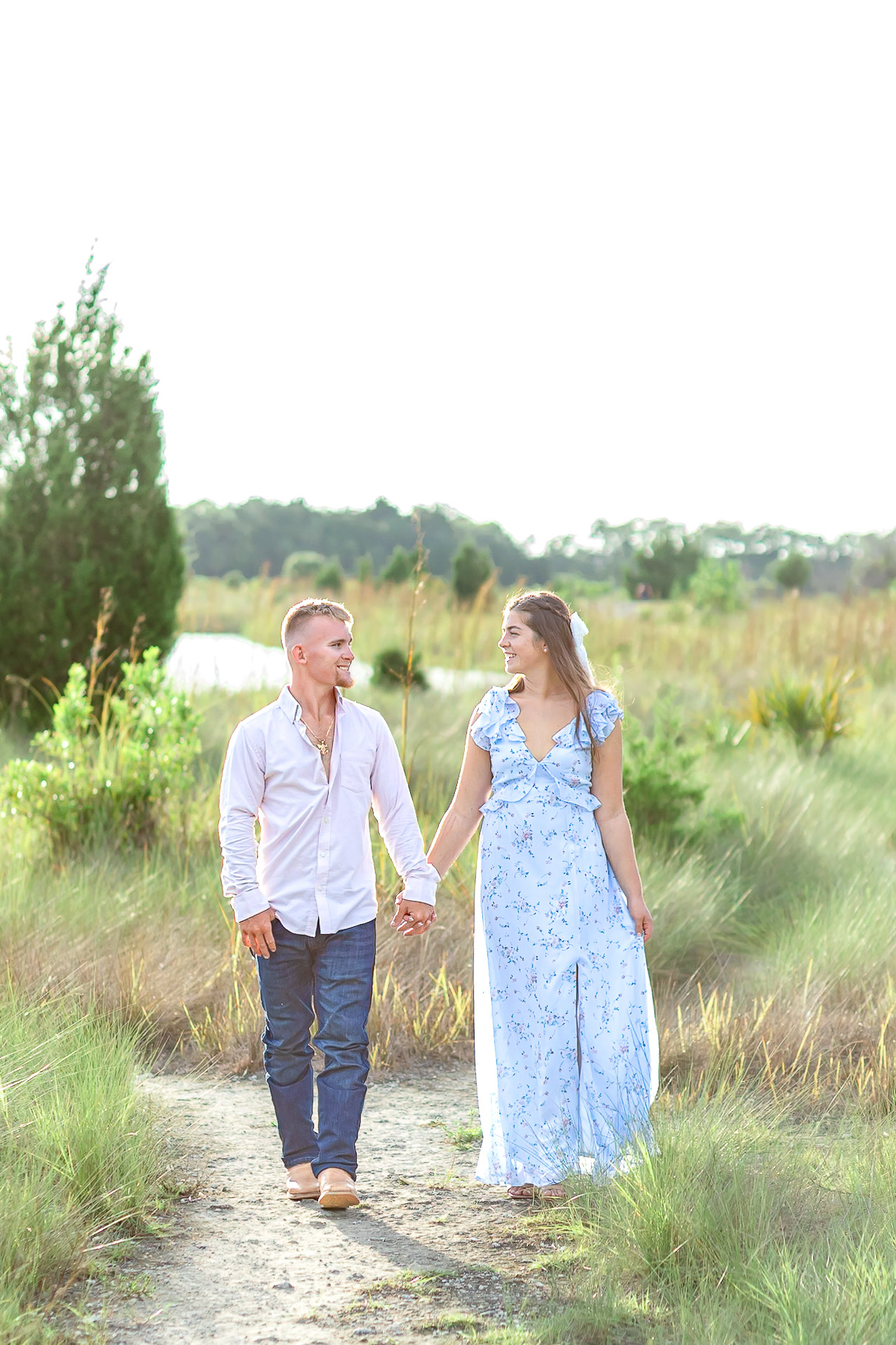 a couple walking on a white trail, surrounded by tall grass. They are dressed up and looking at each other, lovingly,and smiling.