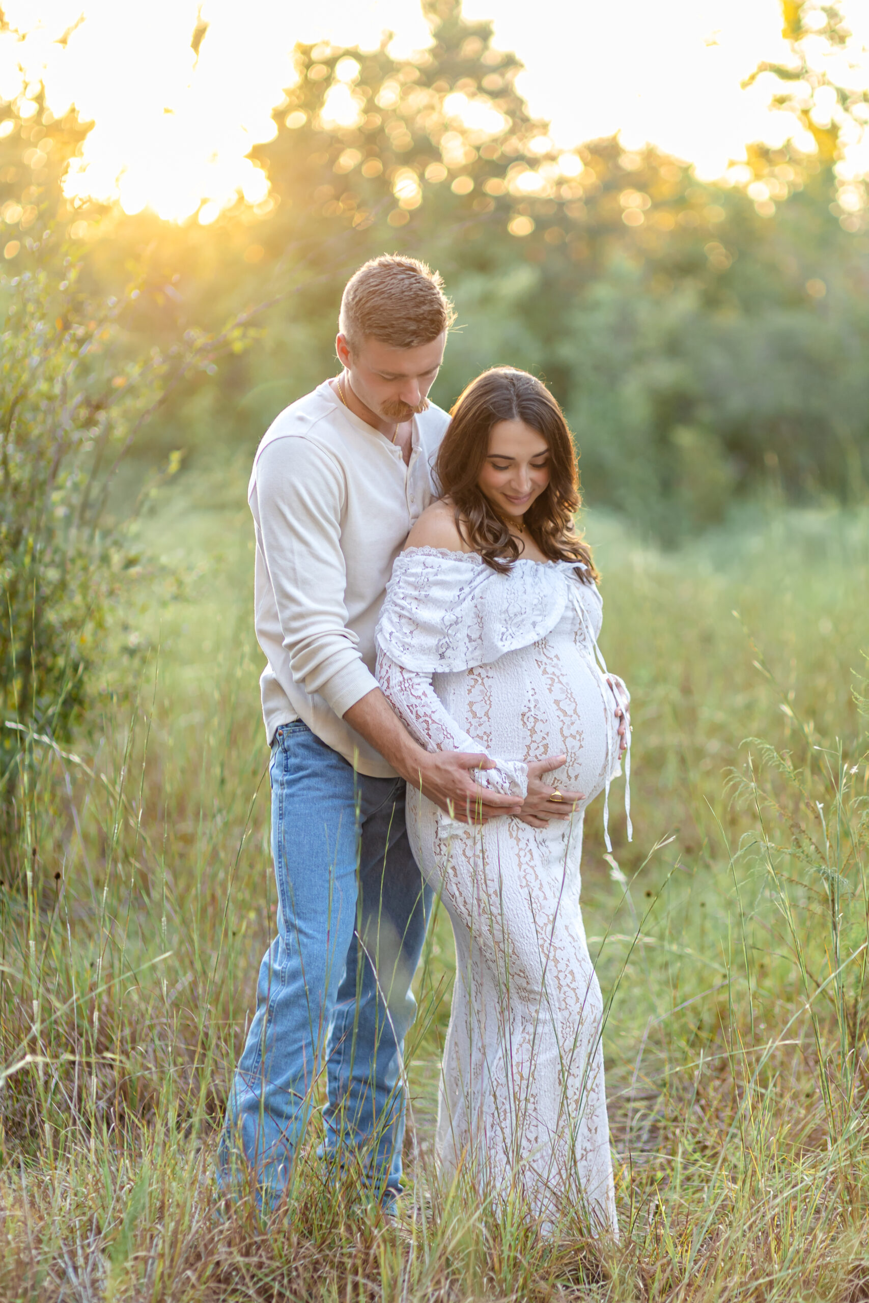 A husband and wife, in a field at sunset, looking at the pregnant tummy of the mom to be.